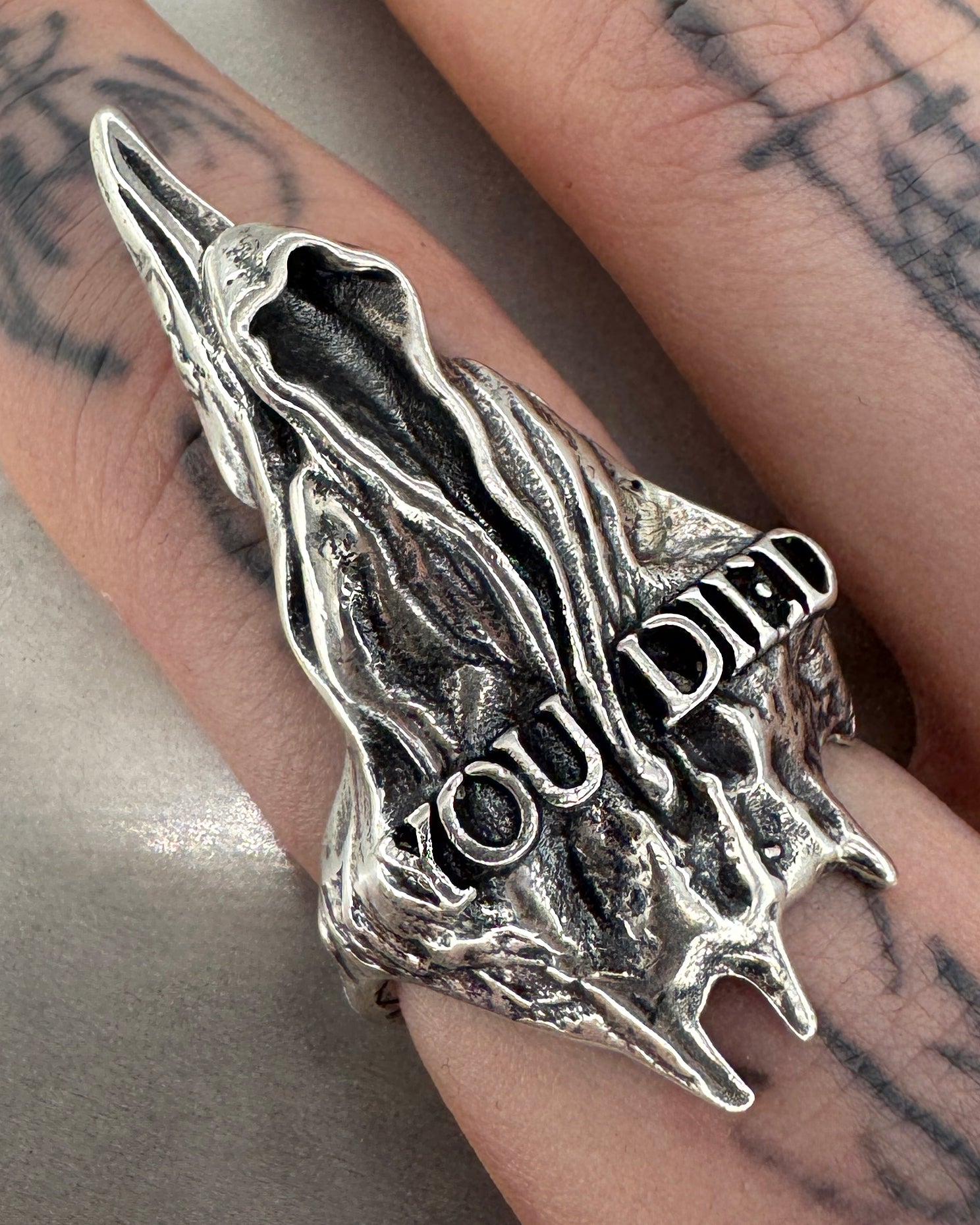 A silver ring with an oxidized finish featuring a hooded figure design with the text 'YOU DIED' engraved on it, on a male figures hand with tattoos.